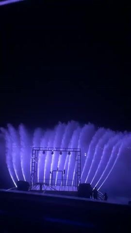 A water show is in progress at night, with water jets illuminated by purple and blue lights. The water is sprayed in a fan-like pattern behind a stage structure. The stage is a metal frame with lights mounted on top. In the distance, city lights are visible on a horizon. Two people are standing on a platform to the right of the stage. The sky is dark.