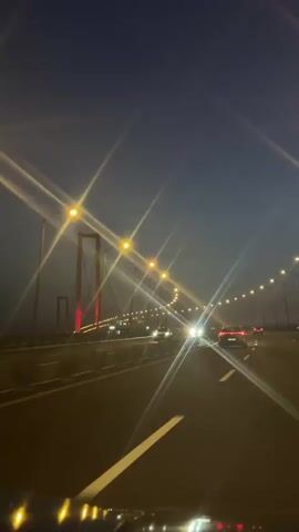 A car travels down a highway bridge at night. Overhead lights illuminate the road. A large suspension bridge tower with red lights is visible in the distance. Other vehicles move in the same direction. The sky above is dark blue.