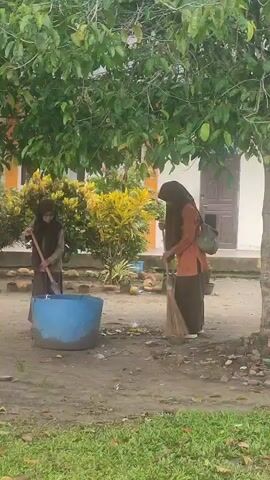 Two women wearing headscarves are cleaning a outdoor area. One woman in an orange top and brown skirt sweeps the ground with a broom. The other woman in a brown dress and brown headscarf stirs something in a large blue tub with a stick. There is a tree to the left and a building in the background.