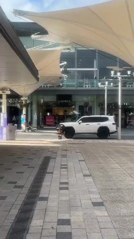 A white SUV is parked on a paved walkway in front of a Gazman clothing store. The store has large windows displaying merchandise and sale signs that read "50% OFF". Several people are visible inside and outside the store. The walkway is lined with shops and covered by a large, white fabric awning. The weather appears to be sunny.