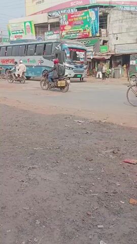 The video depicts a street scene in what appears to be a bustling town or city in South Asia. In the foreground, there's a rough, unpaved area with scattered debris. Several people are riding motorcycles and bicycles along the road. A man pulls a cart attached to a bicycle. A large blue and silver bus is prominent in the background, and billboards with images and text are visible above the buildings. The weather seems overcast, suggesting a cloudy day. Overall, the scene portrays everyday life and transportation in a busy urban environment.