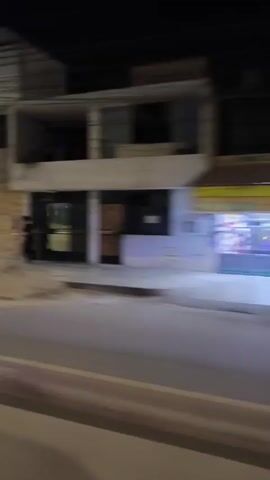 The video depicts a street scene at night, filmed from a moving vehicle. Buildings line the street, some with visible signage and lit windows. A person is seen running along the sidewalk in the initial frames. Cars are visible on the road, and streetlights illuminate the scene. The camera movement causes a blur of the surroundings, but the essential elements of a city street at night are present.