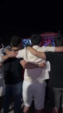 Okay, here’s a description of the video:

The video shows four young men with their arms around each other, facing a stage at what appears to be an outdoor concert or festival. The men are positioned in a line, with the camera behind them, providing a view of their backs. One man is wearing a white shirt with white pants, one is wearing a black shirt and blue jeans, one is wearing a black shirt and khaki pants, and one is wearing a light blue shirt and light colored pants. The stage is in the background, illuminated with lights. The stage has a red banner at the top, with writing in white and a Turkish flag. The night is dark. Other people can be seen in the crowd behind them.