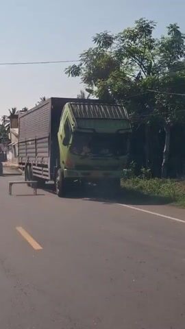 A green truck with its hood up is parked on the side of a road, partially blocking traffic. A dark-colored cargo container is attached to the truck. On the road in front of the truck, a white bench is placed. Further down the road, a red plastic barrier is visible. A person on a motorcycle with a sidecar is seen driving past the barrier. The scene appears to be in a rural or suburban area with houses and trees visible in the background under a clear sky.