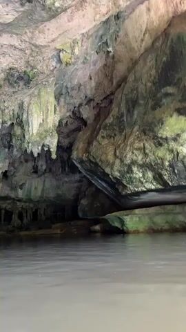 The video shows a body of water with ripples reflecting light. Above the water, a cave ceiling is visible, adorned with stalactites. The cave walls are rough and textured, with patches of green moss or algae. The overall scene suggests an underwater or water-level view within a cave.