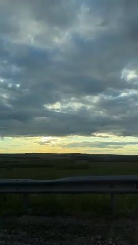 The video shows a view of a landscape from a moving vehicle. In the foreground, there is a metal guardrail and some grass and gravel. The midground consists of a green field with scattered trees. The background features a wide, expansive sky filled with dark, dramatic clouds. Sunlight breaks through the clouds in the distance, creating a bright horizon with a golden hue. The overall impression is of a somewhat overcast but beautiful natural setting, possibly during sunrise or sunset, viewed from a roadside perspective.