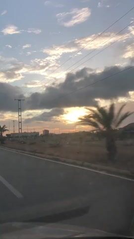 A video captured from a moving vehicle shows a sunset over a rural landscape. The sky is filled with a mix of dark and light clouds, with the sun partially obscured by them, casting a golden glow. Power lines stretch across the frame, supported by tall poles. The road ahead is asphalt with white lane markings. On the left side of the road, there is a lone palm tree and some sparse vegetation. On the right, there is a barren landscape with some scattered bushes and distant hills. The overall impression is one of travel during a scenic dusk.