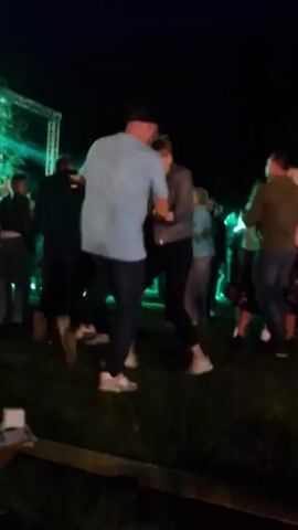 The video shows a night-time outdoor gathering with a group of people dancing. In the foreground, a man in a blue t-shirt and jeans, wearing a baseball cap, is dancing with a woman in a grey jacket and black pants. They are holding hands and appear to be engaged in a lively dance. Several other people are visible in the background, also dancing and socializing. The scene is illuminated by green and white stage lights, and there is a stage structure with lights in the upper left corner of the frame. The ground appears to be covered in grass, and there is a wooden structure or bench in the lower part of the frame. The overall atmosphere suggests a festive event or party.