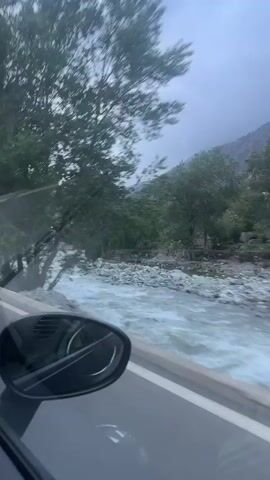 The video captures a first-person perspective from a moving vehicle, looking out the driver's side window. The car's side mirror is prominently visible in the lower left foreground. A wide, rapidly flowing river, with turbulent and milky-colored water, runs parallel to the road in the middle ground.

Numerous green trees line the riverbanks and extend into the mid-distance. Beyond the trees, rugged mountains or large hills are visible under an overcast, grey sky, suggesting cloudy weather or recent rainfall which contributes to the river's high and fast-flowing state. Brief glimpses of a small bridge and some structures or buildings can be seen across the river through the foliage. The continuous motion indicates travel through a scenic, mountainous landscape.

This scene depicts a drive through the natural environment of Asgaour, Morocco, characterized by its powerful river, dense vegetation, and mountainous terrain. There is no visible text in the video.