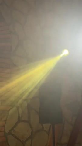 A spotlight beams yellow light across a smoky room. The light emanates from a fixture mounted high on a wall, the light source itself is a bright yellow circle. The beam of light is wide and illuminates the smoke, creating a visible shaft of light. The room appears to have a stone wall construction with brick accents. A black speaker on a stand is visible in the lower right quadrant of the frame. The overall atmosphere suggests a dimly lit venue, possibly a club or event space, with atmospheric lighting effects.