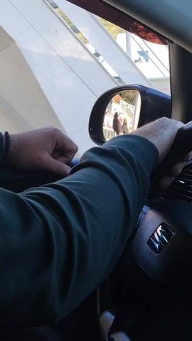 The video shows a person sitting in the driver's seat of a car, with their hands on the steering wheel. The person is wearing a long-sleeved green shirt and has a bracelet on their wrist. The car's side mirror shows two people standing outside, seemingly engaged in a conversation. The environment outside appears to be a daytime setting with buildings visible.