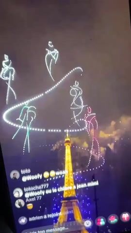 This is a screenshot of a vertical video, likely from a social media platform, showing a nighttime drone show above the Eiffel Tower in Paris. The Eiffel Tower is illuminated in yellow. Drones, forming patterns of lights, are visible in the sky, resembling stylized human figures in motion and a large, swirling loop. The drone figures are depicted in white, light blue, and pink. A comment section is overlaid on the bottom of the screen, showing usernames such as "toto," "totoche777," and "Axel 77," along with brief comments and emojis. The overall environment is dark, indicating nighttime, with some cloudy sky visible.