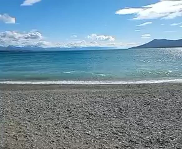 The video shows a pebble beach on a bright sunny day.  Calm, turquoise water is visible, with small waves gently reaching the shore. In the distance, there is a landmass or island on the right, and snow-capped mountains are seen on the left horizon.  The sky is blue with scattered white clouds. The view is static and provides an overall impression of tranquility.
