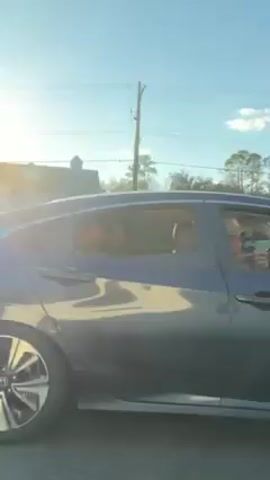 A blue car drives past the camera, with the sun shining in the background. A man wearing a hat is visible in the driver's seat.