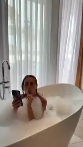 A woman with long brown hair is sitting in a white, egg-shaped bathtub filled with bubbles. She is holding a smartphone with a clear case and a sticker of lips on it. She is looking at the phone and touching her nose. Behind her, there is a large window with sheer white curtains and a view of trees outside. The bathtub has a modern-style chrome faucet with two handles. The bathroom appears to be spacious and well-lit.
