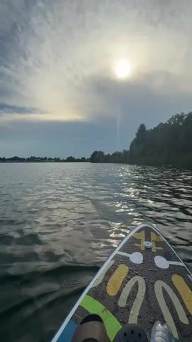 The video opens with a low-angle shot of a body of water, likely a lake or calm sea, from the perspective of someone on a paddleboard. The sun is visible in the upper right quadrant of the frame, partially obscured by clouds, casting a bright glare on the water. The water's surface is rippled, reflecting the sunlight and the sky. The paddleboard itself is visible in the foreground, occupying the bottom right portion of the frame. It has a colorful, graphic design on its surface, and what appears to be a shoe or foot is visible at the very bottom edge. The background shows a distant shoreline with trees and hints of buildings. The sky is a mix of blue and white clouds, with the sun creating a bright halo effect. The camera is positioned just above the water, giving a sense of movement and immersion. The overall impression is one of a serene outdoor activity on a sunny day.