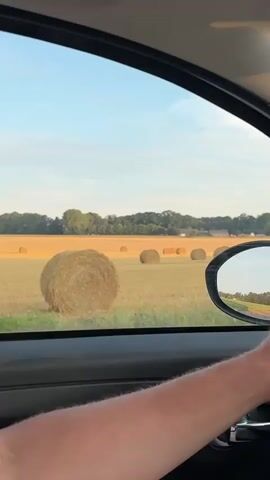 The video shows a view from inside a car, looking out the passenger side window. The car is moving. The landscape outside consists of a field with several round hay bales. In the distance, there is a line of trees and a building. The sky is blue with a few wispy clouds. The sun is low in the sky, suggesting it is either early morning or late afternoon. A person's arm and hand are visible on the steering wheel, indicating they are driving. A side-view mirror reflects the passing scenery. The interior of the car is dark, with the door panel and window frame visible.