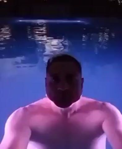 A person with dark hair and a light-skinned complexion is in a pool, with the water illuminated in blue and pink. They appear to be treading water, holding themselves upright, and staring directly at the camera. Light glints off the surface of the water, creating subtle ripples. The person's face is partially submerged. The background is dark, possibly indicating nighttime.