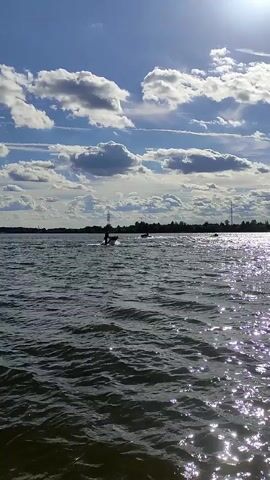 A jet ski is seen from the side as it moves across a body of water, leaving a spray behind it. The sky is blue with white clouds and the sun is shining, creating glints on the water. A treeline is visible in the distance.