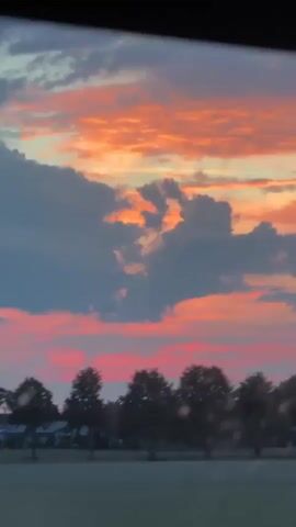 The video shows a sunset sky with orange and pink clouds. There are trees and houses in the background. The video is blurry and appears to be shot from a moving vehicle. There are no people or distinct objects visible.