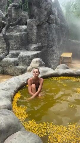 A young woman in a black swimsuit is sitting in a round hot tub surrounded by gray rocks. The water in the tub is a brownish-yellow color and is covered in small yellow flowers. Behind the tub is a large gray rock formation with several levels and some greenery hanging down. A small bamboo bench sits to the right of the rock formation. Steam rises in the background, obscuring the view. The woman looks to her left, then turns to face the camera and smiles briefly.  She then looks down and begins to move in the water.