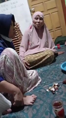 The video shows a group of people sitting on the floor, which is covered with a patterned blanket. There are several plates of fruit, including salak and apples, scattered around. Some of the fruit appears to have been peeled. There are also cups of drinks and a blue plastic plate. The people are dressed in casual clothing, with some wearing hijabs. The room appears to be a simple interior with wooden doors and walls. One person, a woman wearing a pink hijab and patterned skirt, is speaking animatedly, gesturing with her hands. Another person, a man in a black shirt and green pants, is also visible and appears to be listening. The overall scene suggests a casual gathering or meal.