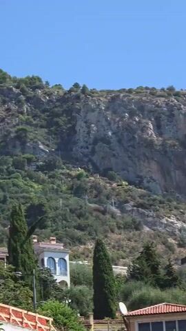 The video shows a view of a large, rocky mountain face. In the foreground, there are several buildings with red tile roofs and palm trees. The sky is clear and blue. The camera appears to be moving slightly to the right, panning across the scene.