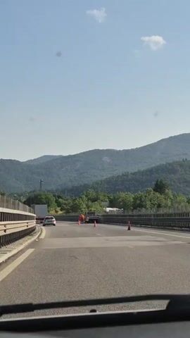 The video shows a highway with construction workers and a tow truck. Two workers in orange vests and pants are standing near the back of a flatbed tow truck. One worker appears to be holding a long pole or tool. Traffic cones are placed on the road around the work area. The truck is on the right side of the road, and there is a guardrail to the left. In the background, there are green, tree-covered mountains under a clear blue sky with a few scattered white clouds. A white car is visible in the distance on the highway. The camera appears to be inside a vehicle moving along the highway.