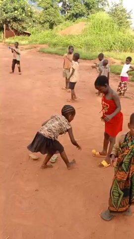 Several young children, predominantly African, play together in an open, dirt-covered area. They are dressed in a variety of casual clothing, some with sandals. One child wears a colorful patterned wrap and manipulates a homemade toy consisting of a stick with attached straws, blowing into it. Another child throws a small, yellow ball. Other children stand around, observing and interacting. The background includes greenery, trees, and a small brick building. The scene is brightly lit, suggesting daytime.