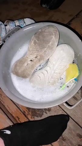 The video shows a top-down view of a gray plastic wash basin filled with soapy water and two shoe soles soaking in it. One sole is a light cream color and appears cleaner, while the other is a darker, dirtier beige with visible tread marks and some debris. To the right of the soles, a yellow sponge and a green and blue toothbrush are submerged in the water. The basin is placed on a weathered wooden surface, possibly a deck or porch. In the foreground, a black sock is visible, with a portion of a bare foot extending into the frame. The lighting suggests it is nighttime or a dimly lit indoor setting.