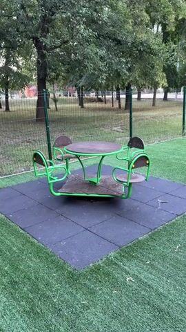 A merry-go-round with four seats and a table in the center sits on a black rubber mat surrounded by artificial turf. It is located in a park-like setting with trees and a fence in the background. The video shows the merry-go-round slowly rotating.