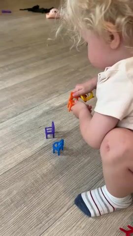A young child with blonde curly hair is playing on a wooden floor. The child is wearing a light-colored t-shirt and striped socks. In front of the child are three small, colorful plastic chairs: one orange, one purple, and one blue. The child is holding a yellow toy in their right hand and is reaching towards the orange chair with their left hand. The child's body is blurred due to motion. The background is a light brown wooden floor with some other small toys scattered around.