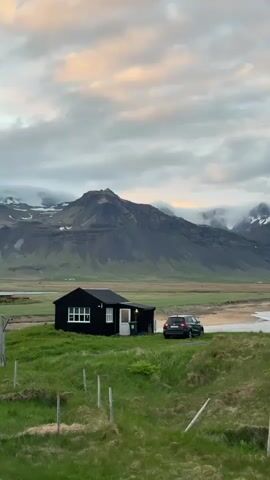 This video captures a panoramic view of a serene coastal landscape in Búðir, Iceland. The camera slowly pans from left to right, revealing a picturesque scene under an overcast sky with hints of soft pink and orange hues, suggesting either sunrise or sunset.

In the foreground, lush green grass covers rolling hills, with a simple wooden fence line visible. To the left, a small, black-painted house with white window frames stands alone, contrasting sharply with the vibrant green surroundings. A dark-colored car, likely an SUV, is parked nearby, facing towards the coast.

Beyond the house and car, a sandy beach stretches along the shore, meeting a calm body of water, likely the ocean or a bay. In the background, majestic mountains dominate the horizon. Many of the rugged peaks are partially obscured by low-hanging clouds, and some higher elevations show patches of snow, even though the foreground is green. The overall impression is one of remote beauty and tranquil wilderness. There is no visible text.