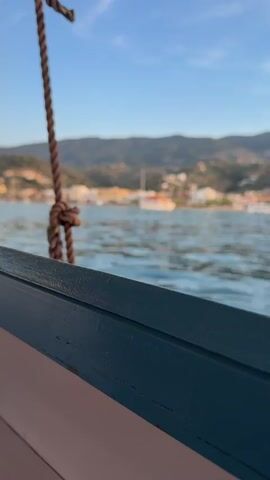 The video opens with a close-up, low-angle shot from a boat, focusing on a dark blue-green wooden boat rail in the foreground. A thick, brown, braided rope, tied with a knot, hangs vertically just behind the rail. The camera then slowly tilts upwards and pans to the right, revealing a serene coastal landscape.

In the midground, a calm body of water stretches across the frame, its surface showing gentle ripples. Across this water, a picturesque town or village is visible, built along the shoreline and extending up the lower slopes of a verdant, mountainous backdrop. The buildings are predominantly white with warm-toned, possibly terracotta, roofs, characteristic of Mediterranean architecture. Several small boats can be seen in the water near the town's waterfront. The mountains behind the town are covered in green vegetation under a bright, clear blue sky with a few wispy clouds.

The lighting suggests late afternoon or early evening, with a warm, soft glow illuminating the scene. The overall atmosphere is peaceful and scenic. The sounds in the video include the gentle lapping of water and indistinct human voices, indicating conversation from people on the boat. No visible text is present. The setting is consistent with the provided context of Poros, Greece.