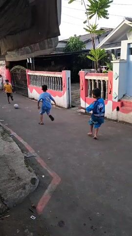 Here is a detailed description of the video:

The video shows a few children playing football (soccer) in a residential street in what appears to be a neighborhood in Southeast Asia. Three children, one in a yellow shirt, and two in matching blue outfits, are directly involved in playing. Another few children are seen observing from a distance in front of a house. The game involves kicking and chasing a small, gray soccer ball. The street is paved and narrow. The environment has houses with fences and some greenery. The general setting suggests a warm climate. The camera is handheld and moves slightly, tracking the movement of the children and the ball.