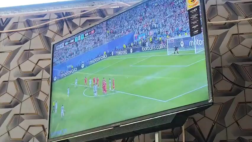 A widescreen television displays a soccer match. The game appears to be in progress, with players on the field and a large crowd in the stadium seating. The screen is mostly filled with the green soccer field and the players in their uniforms. The television is mounted on a stand, and the background wall behind the TV has a patterned wallpaper. The video is a single static shot.
