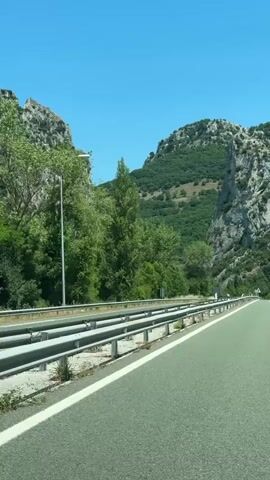 The video captures a view from inside a vehicle driving on a paved road. The road has clear white lane markings and a metal guardrail on the left side. The surrounding environment is mountainous with lush green trees and rocky cliffs. The sky is a clear, bright blue. A few cars are visible on the road ahead. The perspective suggests the vehicle is moving forward.