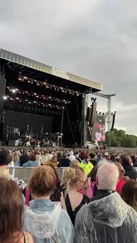 A live outdoor music concert is depicted. A large stage with professional lighting and sound equipment is center frame. A screen above the stage displays a performer. A large crowd of attendees, many wearing ponchos, are visible in the foreground, facing the stage. The sky is overcast. The event appears to be a "Summer Sessions" festival.