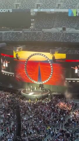 The video shows an aerial view of a concert taking place in a large stadium. The stage is prominently featured with a massive LED screen displaying dynamic visuals. The screen shows a circular graphic superimposed over images of a desert landscape with a road leading towards the horizon, then transitioning to a lush green forest. On the stage, a group of performers are seen on a circular platform. Several people are scattered around the stage, possibly band members or dancers. In front of the stage, a vast crowd of spectators fills the stadium seating, many holding up their phones to record. The stadium itself appears to be a large, open-air structure with tiered seating and large screens visible in the background. The overall atmosphere suggests a live musical performance with elaborate stage production and a large audience.