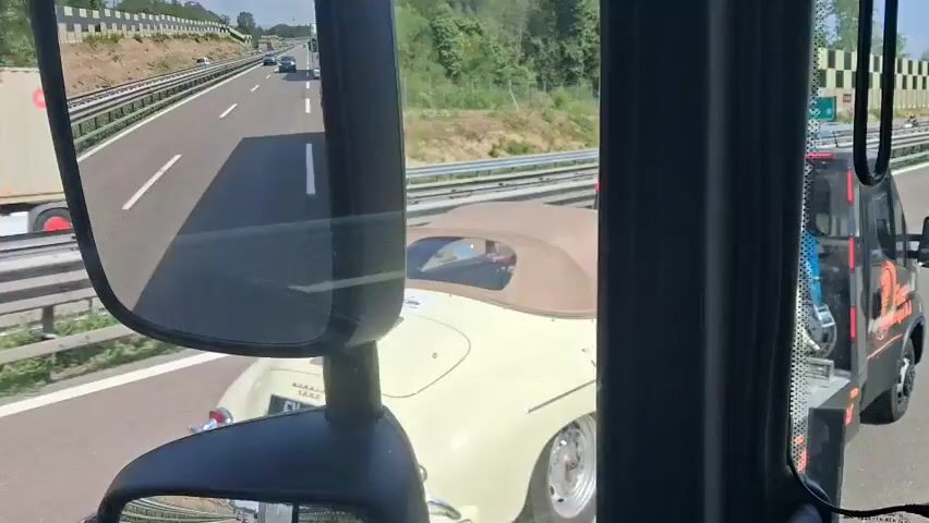 A vintage white convertible Porsche is being transported on a flatbed tow truck on a highway. The car has a tan convertible top. The tow truck is black with orange accents. The license plate on the Porsche reads "FV 186-TD". The truck is driving on the right side of the highway. There are other cars and trucks on the highway in the distance. The environment appears to be sunny with a roadside barrier and some trees visible.