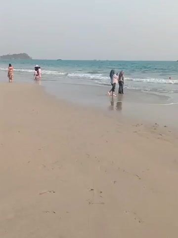 A wide shot captures a beach scene with the ocean in the background. The sky is a pale blue, indicating daylight. Several people are present on the wet sand near the water's edge. In the mid-ground, two individuals, possibly young women, are interacting. One is wearing dark clothing, and the other is wearing lighter clothes and a headscarf. They appear to be engaged in conversation or posing for a photo. To the left, further down the beach, another person is walking away from the camera. In the distance, small boats or ships are visible on the horizon. The waves are gentle, creating a wet sheen on the sand where they have receded. The sand itself is light brown and appears relatively smooth, with some footprints visible. The overall atmosphere is peaceful and suggests a casual outing at the beach.