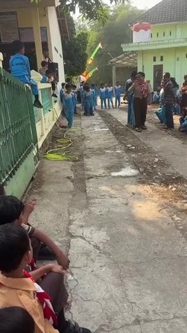 A group of children, mostly wearing blue uniforms, are participating in an outdoor activity. Some of the children are dressed in scout uniforms. The children are arranged in lines, facing each other down a narrow paved path. One group of children is standing on a long, flat object that appears to be a wooden plank, and they are working together to move it along the path, with some children walking on the plank and others pushing or pulling it from the sides. Other children are lined up on either side of the path, observing the activity. There are buildings visible in the background, and trees and foliage suggest a rural or semi-rural setting. The sun is shining, casting shadows, and the overall atmosphere appears to be one of an organized game or competition.