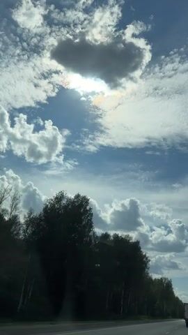 This video shows a view from inside a vehicle traveling on a highway. The sky is partly cloudy with the sun peeking through a large cloud, creating a halo effect. On the left side of the road, there is a dense line of trees. On the right side, more trees line the road, and a road sign is visible. In the distance, a white truck is driving in the same direction as the camera. The road appears to be a two-lane highway with a clear shoulder. The overall impression is of a scenic drive through a wooded area on a sunny day.