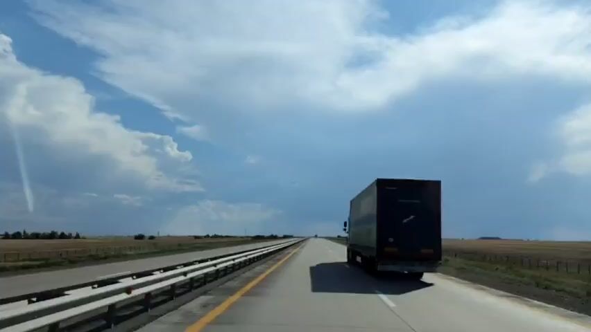 The video shows a view from inside a vehicle traveling on a highway. A large semi-truck with a dark trailer is visible in the right lane, ahead of the camera. The trailer has "EXPEDITION" written on the back. The highway stretches into the distance, with fields on either side. The sky is blue with large, white and dark clouds.