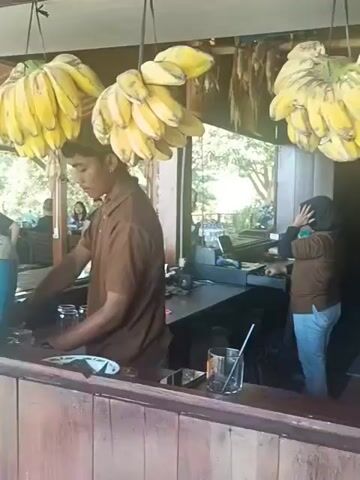 This is a medium shot video taken from the perspective of someone standing in front of a wooden counter. In the foreground, the wooden counter takes up the bottom portion of the frame. Hanging from above the counter are several bunches of ripe yellow bananas. Behind the counter, a person wearing a brown short-sleeved shirt and a dark head covering is preparing drinks. To the left of this person is a large, blue and beige water cooler. In the background, other people are seated at tables, and trees and foliage are visible through an open-air structure. On the right side of the counter, another person wearing a brown shirt and a headscarf is also working. The overall scene appears to be an outdoor cafe or restaurant.