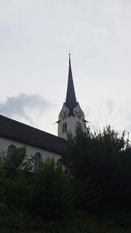 A static medium shot captures a church steeple and part of the church building. The sky is overcast and light gray. The church is white with a dark gray tiled roof. A tall, dark gray steeple with a spire points towards the sky. The steeple has two round clock faces, each with Roman numerals and red clock hands. The time appears to be approximately 9:40 or 9:41. Lush green trees and foliage are visible in the foreground and partially obscure the church building. A dark gray concrete structure is visible at the very bottom of the frame. There are no people visible. The video appears to be a short, unedited clip.