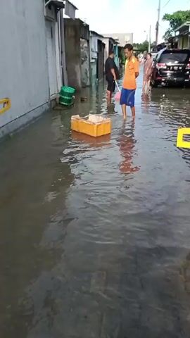 The video shows a flooded street in what appears to be a residential area. The water level is high enough to cover the road surface, reaching above the ankles of the people walking through it. There are several people wading through the water, including two men in the foreground. One man is wearing an orange shirt and blue shorts, and the other is wearing a black shirt and shorts and carrying a pink bucket.  A wooden crate floats in the water. In the background, there is a black SUV parked in the flooded street. Houses and buildings line both sides of the street, and power lines are visible overhead. The sky is partly cloudy.
