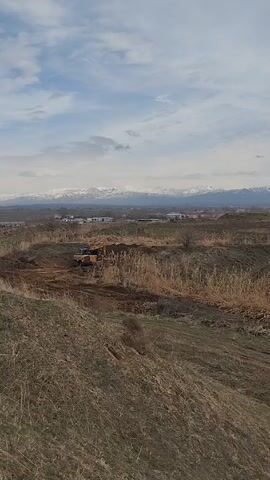 A yellow and black excavator is working in a dirt field. The field is overgrown with dry, brown grass and weeds. In the background is a small town or industrial area. Beyond that are snow-covered mountains under a cloudy sky.