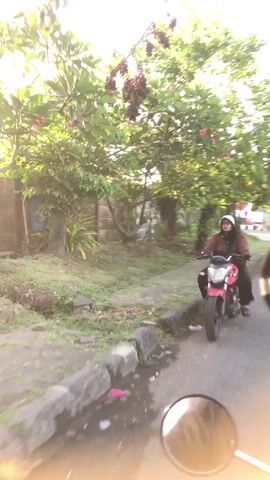 A person wearing a hijab and a maroon jacket rides a maroon motorcycle on a paved path. The motorcycle is moving to the right of the frame. The path is bordered by a curb and a grassy area with some scattered stones. Behind the path is a stone wall and several trees with green leaves and some dried branches. Sunlight filters through the trees, creating a bright, dappled effect. The foreground of the image shows a blurred reflection of a motorcycle's rearview mirror, suggesting the video is being filmed from another motorcycle. The overall environment appears to be an outdoor, possibly urban or suburban, setting during daylight hours.