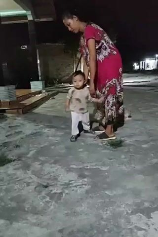 A woman wearing a floral dress stands next to a toddler in a light-colored shirt and pants. The child walks away from the camera. The scene appears to be an outdoor courtyard at night, with a building in the background.
